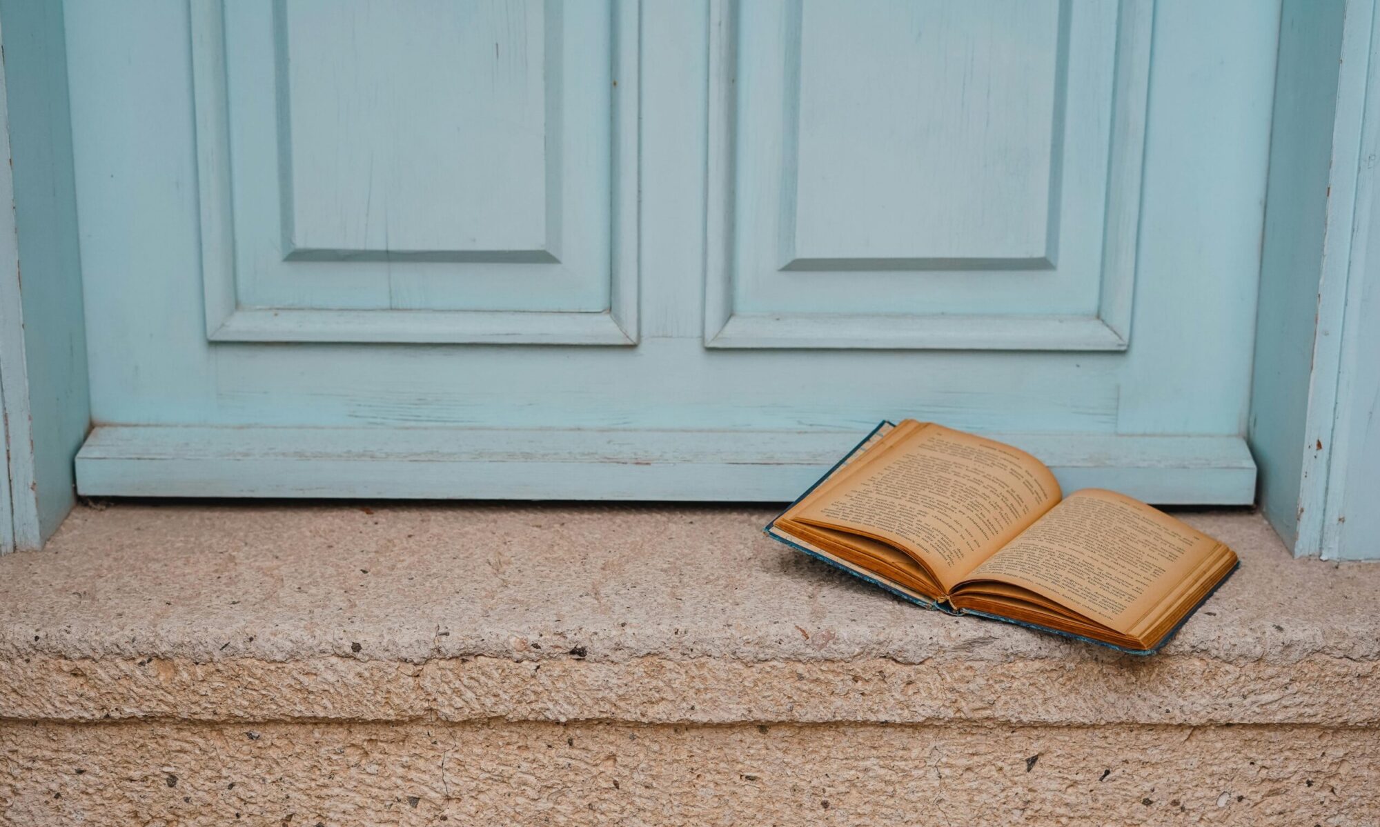 An open book rests on a rustic doorstep with a vintage blue door backdrop.